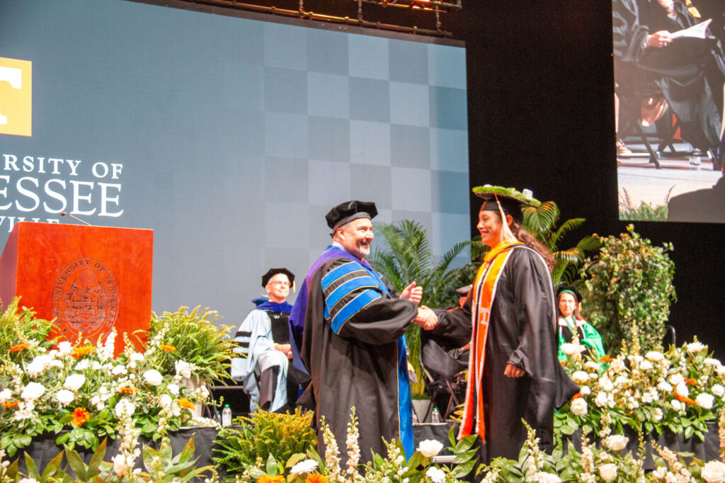 A master's graduate with a grass diorama decorating their mortarboard shakes the hand of the Dean of the Herbert College of Agriculture, David White.